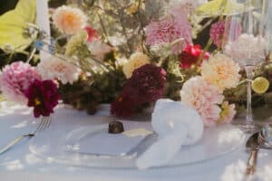 Ein wunderschön arrangierter Tisch, perfekt für eine Hochzeit, mit einem durchsichtigen Glasteller, einer weißen Serviette, einer kleinen Praline und einer Karte. Im Hintergrund leuchtet ein leuchtender Strauß rosa, roter und gelber Blumen sanft im Sonnenlicht.