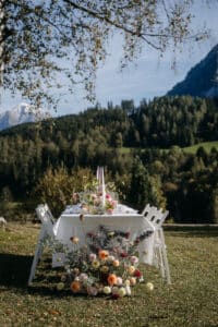 Ein wunderschön gedeckter Esstisch im Freien mit weißen Stühlen ist mit Blumenarrangements geschmückt und bietet eine idyllische Kulisse für eine Hochzeit. Umgeben von üppigem Grün bilden Berge und Bäume eine atemberaubende Kulisse unter einem klaren blauen Himmel, perfekt für eine Hochzeit inmitten der Pracht der Natur.