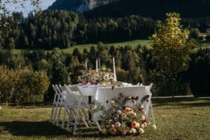 Ein wunderschön gedeckter Gartentisch mit weißen Stühlen und einem Blumenschmuck steht auf einer Wiese, perfekt für eine Hochzeit, umgeben von üppigen grünen Bäumen und sanften Hügeln unter einem klaren Himmel.