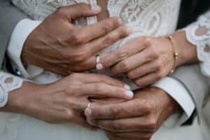 Eine Nahaufnahme zeigt die mit Eheringen geschmückten Hände des Paares. Die Person im Anzug umarmt sich zärtlich von hinten, während die Person im Spitzenkleid zärtlich die Hände ihres Partners hält und so eine perfekte Szene für ihre Hochzeit schafft.