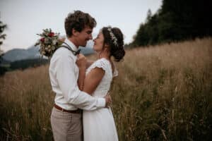 Auf einer malerischen Wiese feiert ein fröhliches Paar seine Hochzeit, während der Mann verspielt einen Blumenstrauß über der Schulter hält. In eleganter Hochzeitskleidung stehen sie inmitten von Graswiesen mit Bäumen und Bergen in der Ferne und verkörpern die Freude, die mit einer Hochzeit einhergeht.