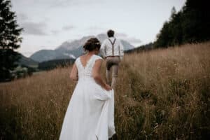 Eine Braut im weißen Kleid und ein Bräutigam in Hosenträgern gehen während ihrer Hochzeit einen grasbewachsenen Hügel hinauf, umgeben von hohen Bäumen, mit Bergen im Hintergrund unter einem bewölkten Himmel.