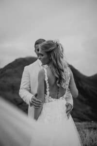 Ein Schwarzweißfoto zeigt ein Paar, das sich im Freien umarmt. Beide sind in formeller Kleidung. Die Frau trägt ein rückenfreies Spitzenbrautkleid, perfekt für eine Hochzeit, während der Mann einen hellen Anzug trägt. Berge zieren den Hintergrund unter einem bewölkten Himmel.