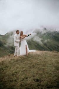 Ein Paar im Hochzeitskleid feiert seine Hochzeit auf einem grasbedeckten Hügel. Die Braut lässt freudig Champagner in die Luft strömen, umgeben von nebligen Bergen und bewölktem Himmel.