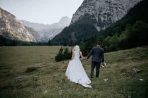Braut und Bräutigam gehen Hand in Hand durch eine Wiese mit einer Bergkulisse im Hintergrund und verkörpern so die Romantik ihrer Hochzeit. Die Braut trägt ein fließendes weißes Kleid und einen Schleier, während der Bräutigam einen grauen Anzug trägt. Sie halten einen ruhigen Moment fest, als sie von der Kamera weggehen.