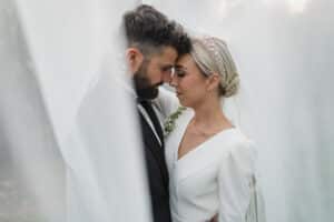 Ein Paar im Hochzeitskleid umarmt sich zärtlich, umgeben von einem weichen, durchscheinenden Schleier. Der Bräutigam trägt einen dunklen Anzug und die Braut ein weißes Kleid mit einem juwelenbesetzten Stirnband. Während dieses intimen Augenblicks sind Andeutungen der Hochzeitsplanung deutlich zu erkennen, da sie eng beieinander stehen und die Augen geschlossen haben.