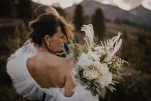 Eine Braut in einem weißen Kleid hält einen Strauß weißer Blumen und Grünpflanzen in der Hand. Mit elegant gestyltem Haar steht sie vor einer Landschaft aus Hügeln und Bäumen unter einem bewölkten Himmel und träumt von perfekten Momenten aus der sorgfältigen Hochzeitsplanung, die sie hierher gebracht hat.