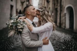 Ein Paar umarmt sich auf einer Kopfsteinpflasterstraße und verkörpert die Essenz einer perfekten Hochzeit. Der Bräutigam küsst zärtlich die Stirn der Braut, während sie einen Strauß weißer und grüner Blumen hält. Beide sind in Abendgarderobe gekleidet. Die Braut glänzt in ihrem Spitzenkleid, während der Bräutigam in seinem hellen Anzug elegant aussieht.