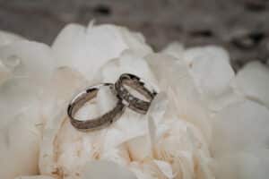 Zwei silberne Eheringe mit komplizierten Mustern ruhen auf weichen, weißen Pfingstrosenblättern und schaffen eine elegante und romantische Hochzeitsplanungsszene.