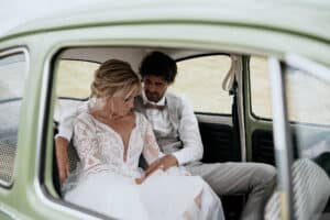 Ein Paar sitzt auf dem Rücksitz eines grünen Oldtimers und verkörpert die Essenz einer perfekten Hochzeit. Die Frau in einem weißen Spitzenbrautkleid lehnt sich liebevoll an den Mann, der ein weißes Hemd und hellgraue Hosen trägt. Die Szene ist heiter und intim.