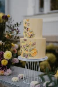 Eine zweistöckige gelbe Torte mit farbenfrohen Blumendekorationen, perfekt für eine rustikale Hochzeit. Im Freien auf einem weißen Ständer präsentiert und von einer Vielzahl von Blumen umgeben, schafft sie eine elegante Atmosphäre, ideal für diejenigen, die schon weit in ihrer Hochzeitsplanung sind.