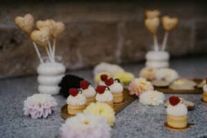 Eine bezaubernde Hochzeitsszene entfaltet sich mit einer Präsentation kleiner Cupcakes mit Sahne und Himbeeren auf einer Steinoberfläche, umgeben von pastellfarbenen Blumen. Herzförmige Kekse auf Stöcken stehen in weißen Vasen und fangen nahtlos die Essenz einer durchdachten Hochzeitsplanung ein.