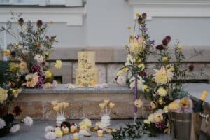 Eine wunderschön arrangierte Hochzeitskulisse im Freien mit einer Torte im Blumenmotiv. Die Szene umfasst eine Vielzahl bunter Blumen, brennende Kerzen, Desserts auf kleinen Tellern und einen Eimer voller Flaschen, die eine elegante und festliche Atmosphäre schaffen, die perfekt für Ihre Hochzeitsfeier ist.