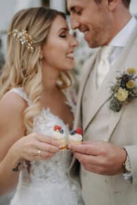 Braut und Bräutigam strahlen sich bei ihrer Hochzeit an und halten kleine Desserts mit Beeren in den Händen. Die Braut trägt ein Spitzenkleid mit einem floralen Haarschmuck, während der Bräutigam einen hellen Anzug mit Ansteckblume trägt.