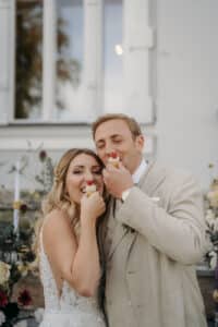 Ein Paar in Hochzeitskleidung teilt einen freudigen Moment, wobei jeder einen Cupcake vor den Mund hält. Sie stehen vor einem hellen Gebäude mit Blumen und Kerzen im Hintergrund, lächeln und genießen die Hochzeitsfeier.