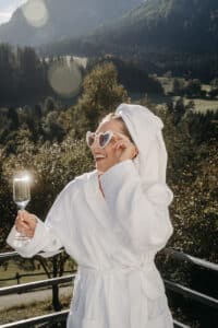 Eine Person im weißen Bademantel und mit Handtuch auf dem Kopf lächelt und hält ein Champagnerglas. Sie trägt eine herzförmige Sonnenbrille und posiert auf einem Balkon mit malerischer Aussicht auf Berge und Bäume und träumt davon, inmitten dieser Schönheit zu heiraten.