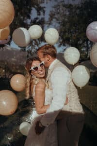 Ein Paar, das sich im Freien umarmt, umhüllt von weißen und goldenen Luftballons, verkörpert den Geist einer Hochzeit. Die Frau glänzt in einem weißen Kleid und herzförmigen Sonnenbrillen, während der Mann in einem hellen Anzug elegant aussieht. Sonnenbeschienene Bäume tragen zur festlichen und romantischen Atmosphäre bei, die perfekt für Hochzeiten ist.