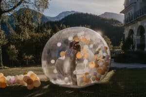 Ein Paar tanzt fröhlich in einer großen transparenten Blase, die mit bunten Luftballons gefüllt ist. Die Kulisse aus majestätischen Bergen, Bäumen und einem bezaubernden Gebäude bietet eine malerische Kulisse für ihre Hochzeit. Wenn die Sonne untergeht, taucht sie diese bezaubernde Feier in einen warmen Glanz.