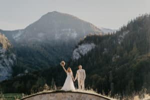 Braut und Bräutigam im Hochzeitskleid stehen auf einer Steinkonstruktion, halten Händchen und Blumen, im Hintergrund sind bewaldete Berge unter einem klaren Himmel zu sehen. In einem Moment purer Freude streckt die Braut ihren Blumenstrauß hoch und feiert ihre perfekte Hochzeit.
