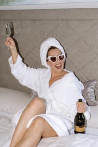 Eine Frau in einem weißen Bademantel und einem Handtuchturban sowie einer herzförmigen Sonnenbrille sitzt mit einer Champagnerflasche in der einen und einem Glas in der anderen Hand auf einem Bett und lächelt energisch – vielleicht feiert sie gerade ihre Hochzeit. Im Hintergrund sind ein gepolstertes Kopfteil und Kissen zu sehen.