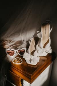 Weiße Pumps mit Schleife auf einem Holztisch, dazu herzförmige Sonnenbrillen mit Perlenrand, eine Perlenkette und Creolen. Im Hintergrund fällt ein zarter Schleier sanft herab – ein perfektes Schaustück für alle, die von ihrer Hochzeit träumen.
