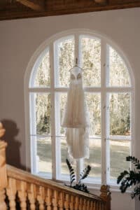 Ein Spitzenbrautkleid hängt vor einem gewölbten Fenster, durch das das Sonnenlicht strömt. Stellen Sie sich bei der Planung Ihrer Traumhochzeit vor, wie Sie die Holztreppe hinauf zu dieser Szene gehen, während das Grün draußen Ihnen eine wunderschöne Reise verspricht.