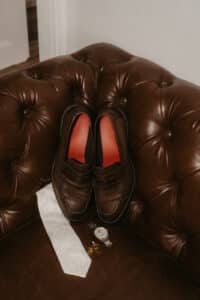 Ein Paar braune Lederhalbschuhe ruht elegant auf einem getufteten braunen Ledersofa, perfekt für jede Hochzeitsplanung. Daneben befinden sich eine weiß gemusterte Krawatte, eine Uhr mit passendem braunen Lederarmband und goldene Manschettenknöpfe, alles ideale Accessoires für den anspruchsvollen Bräutigam, der sich auf seine Hochzeit vorbereitet.