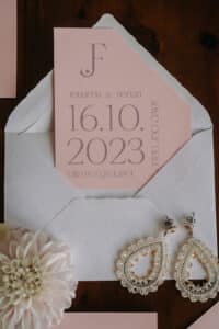 Eine rosa Hochzeitseinladung mit den Initialen „JF“, den Namen „Johanna & Florian“ und dem Datum „16.10.2023“ steckt in einem weißen Umschlag. Daneben liegen Perlenohrringe und eine hellrosa Dahlie, perfekte Details für eine sorgfältig kuratierte Hochzeitsplanungsszene.