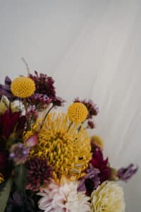 Ein lebendiger Blumenstrauß, perfekt für eine Hochzeit, mit gelben Billy Balls, einer großen gelben Nadelkissen-Protea und verschiedenen violetten, rosa und weißen Blumen vor einem weichen, hellen Hintergrund. Ideal für alle, die sich intensiv mit der Hochzeitsplanung beschäftigen.