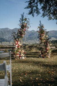 Eine rustikale Hochzeitsszene im Freien mit Blumenbögen, die mit bunten Blumen und Grünpflanzen geschmückt sind. Im Vordergrund sind weiße Stühle aufgestellt, die eine perfekte Kulisse für Hochzeiten bieten. Unter einem klaren Himmel ist eine malerische Aussicht auf Hügel und Berge zu sehen.
