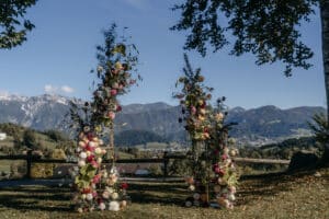 Eine malerische Hochzeitsdekoration im Freien mit zwei verzierten Blumenbögen, die mit bunten Blumen und Grünpflanzen geschmückt sind. Die Kulisse umfasst sanfte Hügel, Berge in der Ferne und einen blauen Himmel im Schatten der Bäume – perfekt für alle, die gerade mitten in der Hochzeitsplanung stecken.