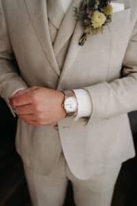 Eine Person im beigen Anzug rückt ihre Armbanduhr zurecht und bereitet sich auf eine Hochzeit vor. Am Revers ist eine Boutonniere mit getrockneten Blumen befestigt. Der Fokus liegt auf den Zeigern und der Uhr, was Eleganz und Liebe zum Detail vermittelt und eine sorgfältige Hochzeitsplanung widerspiegelt.
