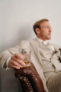 Ein Mann in einem hellen Anzug, der an einen Bräutigam erinnert, der seine Hochzeit plant, sitzt auf einem braunen Lederstuhl und hält ein Glas Whisky in der Hand. Der Hintergrund ist schlicht und er blickt in entspannter Haltung zur Seite, vielleicht denkt er über die bevorstehende Reise nach.