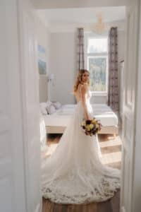 Eine Braut in einem langen weißen Kleid steht in einem sonnendurchfluteten Raum und hält einen Blumenstrauß in der Hand. Sie verkörpert die Essenz einer Hochzeit. Sie blickt zurück zur Kamera, eingerahmt von weißen Türen, einem großen Fenster und weichen Vorhängen, die sanft die Szene für ihren besonderen Moment schaffen.