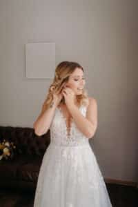 Eine Braut in einem weißen Spitzenbrautkleid, das die Eleganz ihrer Hochzeitsplanung verkörpert, rückt ihren Ohrring zurecht. Ihr langes, welliges Haar fällt herab, während sie lächelt. Im Hintergrund steht ein braunes Sofa mit einem Blumenstrauß und eine leere Leinwand ziert die Wand.