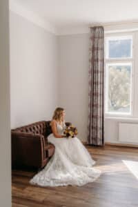 Eine Braut in einem weißen Spitzenkleid, gefangen inmitten ihrer Hochzeitsplanung, sitzt auf einem braunen Ledersofa und hält einen Strauß lila und gelber Blumen in der Hand. Sie blickt aus dem großen Fenster mit gemusterten Vorhängen und denkt über ihre Reise nach Heiraten nach.