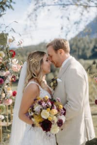 Braut und Bräutigam stehen eng beieinander in einer idyllischen Umgebung im Freien. Die Braut hält einen Strauß bunter Blumen in der Hand, der den Geist ihrer Hochzeit verkörpert. Beide sind elegant gekleidet, umgeben von Blumenschmuck und einer atemberaubenden Berglandschaft, die die Essenz perfekter Hochzeitsplanung einfängt.