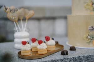 Ein Tablett mit kleinen Vanille-Cupcakes mit Sahne, Himbeeren und Blaubeeren steht neben einer mehrstöckigen Torte mit Blumenschmuck, perfekt für eine Hochzeit. Schokoladentrüffel und herzförmige Cake Pops in einer Vase ergänzen die entzückenden Hochzeitsplanungsdetails.