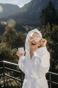 Eine Person steht in einem weißen Gewand und mit herzförmiger Sonnenbrille auf einem Balkon, lächelt breit und hält eine Kaffeetasse in der Hand. Dahinter bietet sich ein malerischer Blick auf üppig grüne Berge und Bäume, die die perfekte Kulisse für romantische Hochzeitsmomente bilden.