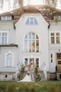 Braut und Bräutigam stehen vor einem eleganten, großen Haus mit Satteldach. Ihre Hochzeit ist von exquisiten Blumenarrangements umgeben. Die Braut hält einen Blumenstrauß, während sie einander anschauen, was eine helle und heitere Szene schafft.