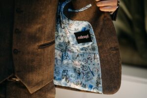 Eine Person hält eine braune Jacke offen, perfekt für eine Hochzeit. Sie zeigt ein blaues Futter mit Abbildungen von Fahrrädern und Reisestempeln. Auf der Innenseite der Jacke steht „Rotknopf“. Der Hintergrund ist unscharf, was diesem exquisiten Stück einen Hauch von Geheimnis verleiht.