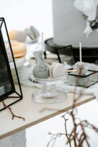 Eine Nahaufnahme einer Dessertpräsentation, die perfekt für eine Hochzeit ist. Sie zeigt ein graues Macaron und einen kugelförmigen Cake Pop mit felsiger Dekoration auf einem kleinen Glasständer. Der elegante Hintergrund ist ideal für die Hochzeitsplanung und präsentiert weiteres Gebäck auf Tabletts und minimalistischem Dekor.