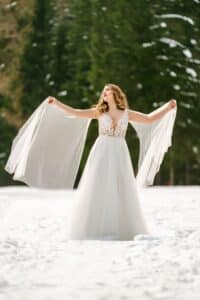 Eine Frau in einem fließenden weißen Brautkleid steht in einer verschneiten Landschaft und verkörpert die Eleganz einer perfekten Hochzeit. Sie hält den hauchdünnen Stoff ihres Kleides wie Flügel ausgebreitet und blickt nach oben, umgeben von schneebedeckten immergrünen Bäumen.