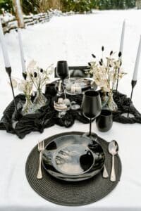 Eine stilvolle Tischdekoration im Freien auf Schnee ist in Schwarz-Weiß gehalten und besteht aus schwarzen Tellern, Gläsern und Besteck. Weiße Blumen und schwarze Kerzen in Kerzenhaltern sind perfekt für eine Winterhochzeit und verleihen der Szene vor einer atemberaubenden Schneelandschaft Eleganz.