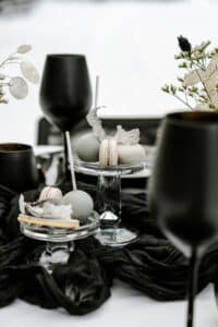 Eine raffinierte Tischdekoration, die perfekt für eine Hochzeit ist, besteht aus grauen Macarons und zarten Süßigkeiten auf Glasständern. Schwarze Weingläser und fließender schwarzer Stoff werten das Arrangement auf, während getrocknete Blumen dem monochromen Thema Eleganz verleihen – ideal für eine sorgfältige Hochzeitsplanung.