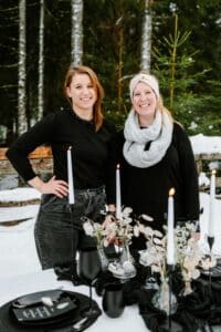 Zwei lächelnde Frauen an einem gedeckten Tisch im Freien in einem verschneiten Wald, perfekt für eine Winterhochzeit. Der Tisch ist mit schwarzen Tellern, weißen Kerzen und dekorativem Grün ausgestattet. Beide sind warm gekleidet, und schneebedeckte Bäume bieten eine zauberhafte Kulisse für ihre Hochzeitsplanung.