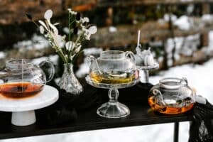 Ein für eine Hochzeit gedeckter Tisch mit drei Glasteekannen mit verschiedenen Teesorten, eine davon elegant auf einem Marmorständer platziert. Eine Vase mit weißen Blumen und Baumwollstängeln sorgt für eine dekorative Note, während der Hintergrund einen verschwommenen, schneebedeckten Waldbereich zeigt.