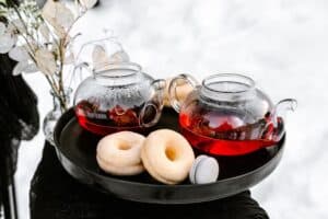 Auf einem schwarzen Tablett stehen zwei Glasteekannen mit rotem Tee, zwei Zuckerdonuts und ein Macaron – perfekt für einen gemütlichen Hochzeitsmoment. Neben dem Tablett steht eine Vase mit zarten, durchscheinenden Blättern. Die verschneite Szene im Hintergrund verleiht dieser intimen Feier eine bezaubernde Note.