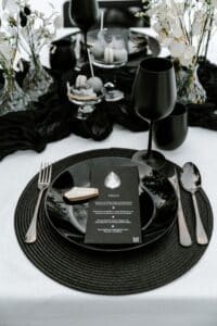 Eine schwarz gedeckte Hochzeitstafel strahlt Eleganz aus mit einem schwarzen Teller, Menü und Tischkarte, ergänzt durch schwarzes Besteck und zwei Kelche. Das Arrangement wird durch Blumenvasen und strukturierte Tischdecken auf Weiß veredelt. Ideal für jede Hochzeitsplanung, die nach Raffinesse strebt.