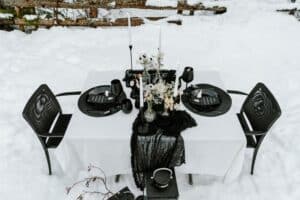 In einer schneebedeckten Außenkulisse, die sich perfekt für eine Hochzeit eignet, ist ein quadratischer Tisch mit einer weißen Tischdecke und schwarzen Stühlen geschmückt. Das elegante Arrangement besteht aus schwarzen Tellern, hohen weißen Kerzen und einem schwarzen Netzläufer, der einen auffälligen Kontrast zum schneebedeckten Hintergrund bildet.